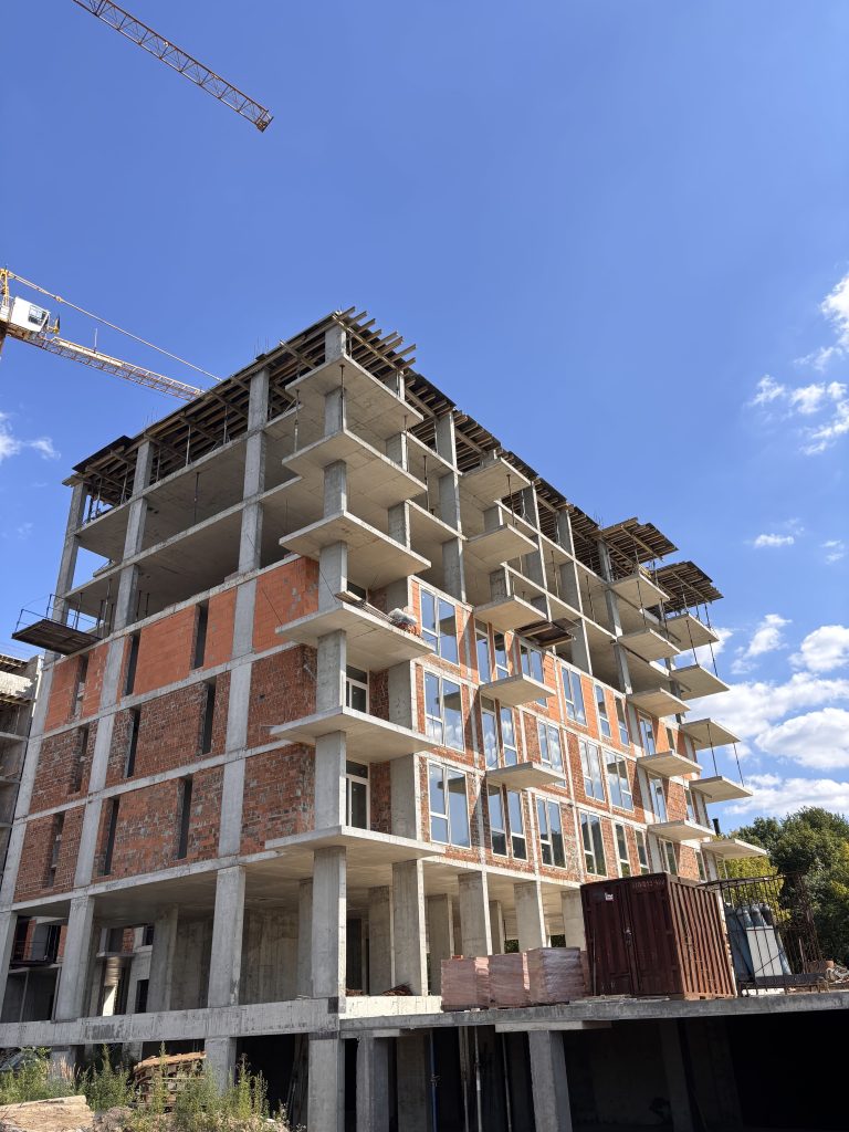 Under-construction concrete and brick building with exposed floors, columns, and scaffolding against a clear blue sky, crane in distance.