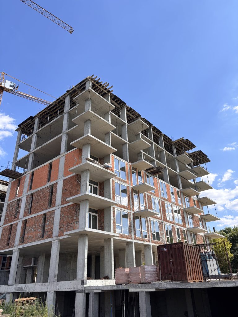 Under-construction multi-story concrete building with brick facade, exposed floors, and a crane in a clear blue sky above.