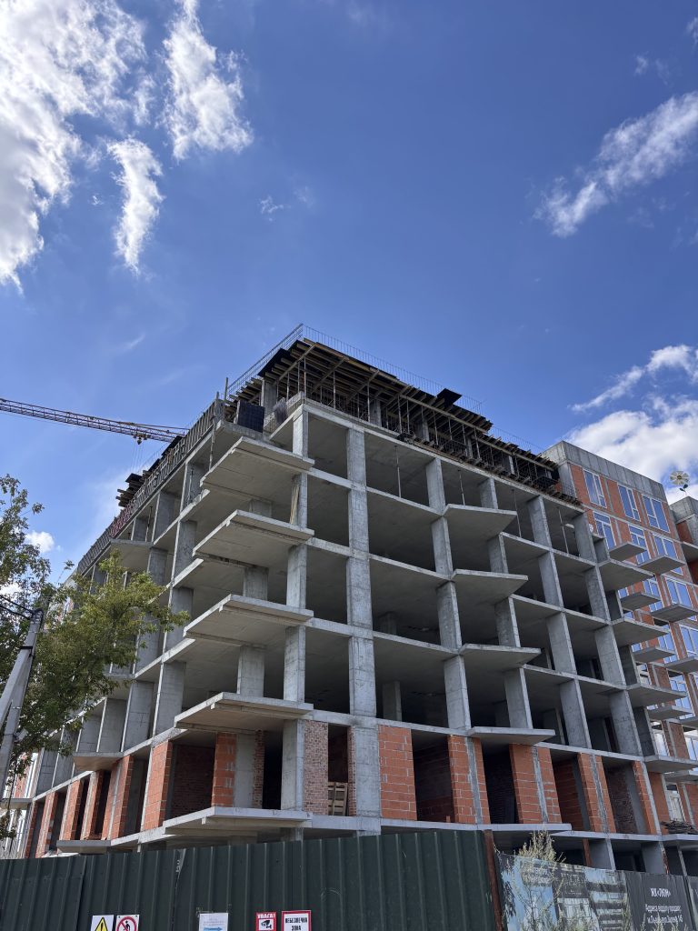 Under-construction concrete building with exposed floors and scaffolding, a crane visible against a blue sky with scattered clouds in the background.