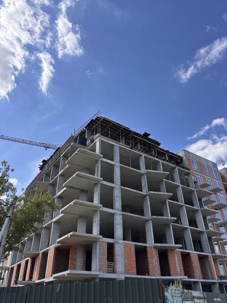 Under-construction concrete building with exposed floors and brick accents against a blue sky with a crane visible in the distance