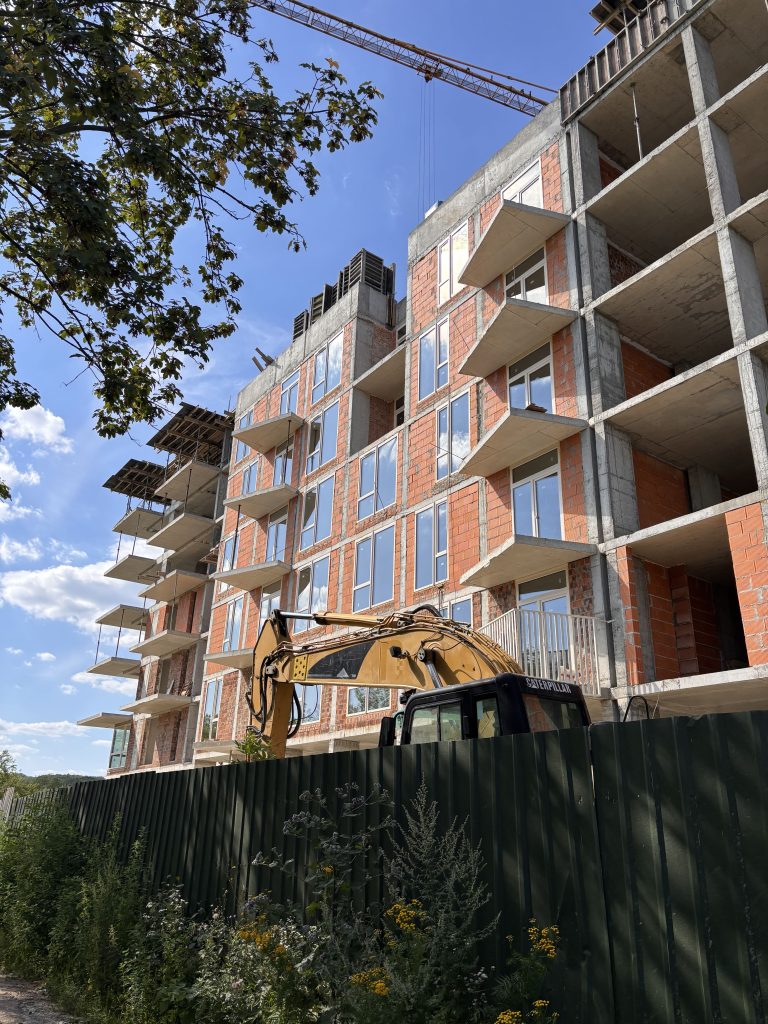 Under-construction multi-story building with exposed brick, concrete frames, and a crane overhead against a blue sky